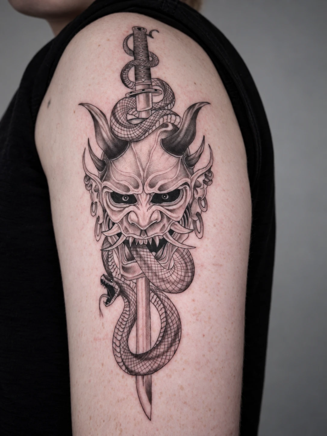 Hannya mask with a katana and a coiling snake, tattooed in black and grey on the upper arm